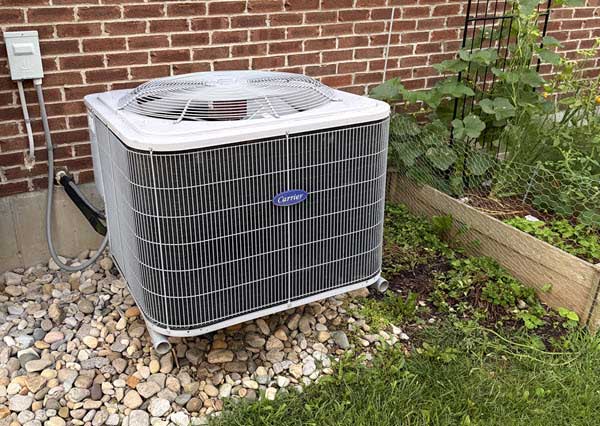 air conditioner | Gardens with Wings Outdoor central air conditioner condenser on gravel beside a brick wall, with a small garden bed nearby.