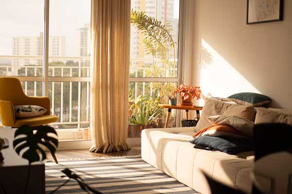 apartment inside | Gardens with Wings Sunlit living room with a beige sofa, mustard chair, and potted plants by a glass balcony door.