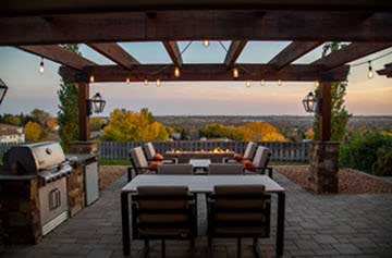 Outdoor patio under a wooden pergola with a built‑in grill, fireplace, and cushioned seating overlooking autumn trees.