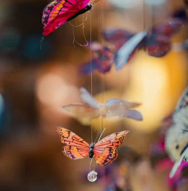 butterfly craft | Gardens with Wings Orange butterfly ornament with black vein patterns suspended on a thin string among colorful blurred decorations in the background.