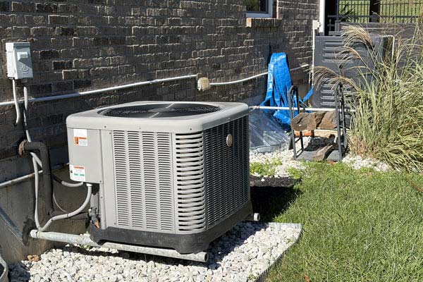 condenser outside | Gardens with Wings Outdoor air conditioning condenser on a gravel pad beside a brick house, with exposed refrigerant lines and a blue tarp nearby.