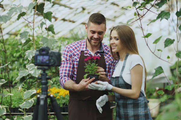 greenhouse flowers | Gardens with Wings Two people in a greenhouse film a gardening video, holding a potted plant in front of a camera on a tripod.