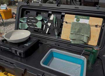 Open rugged equipment case revealing camping kitchen gear: pan, spatula, tongs, green utensils, cutting board with towel, and a blue tub of water.