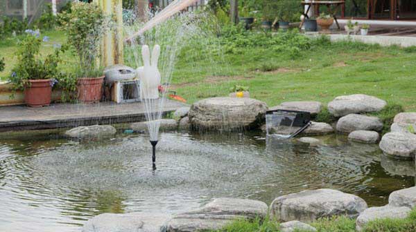 ground fountain | Gardens with Wings Garden pond with a hand-shaped fountain spraying water, surrounded by rocks and greenery.