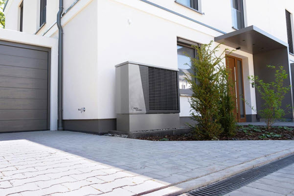 hvac repair | Gardens with Wings Modern white house with a gray garage door, an outdoor HVAC unit on the wall, and a paved driveway with small trees nearby.