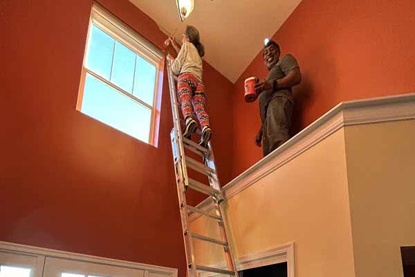 indoor painting | Gardens with Wings Woman on a ladder painting a tall red interior wall; man on the balcony holds a paint can nearby by a window.