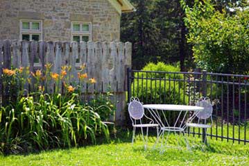 White metal bistro table and two chairs on a sunny lawn, with flowers and a wooden fence in the background.