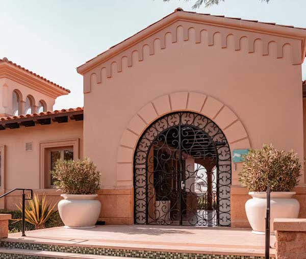 large post | Gardens with Wings Front entrance of a peach Mediterranean-style villa with a tall arched doorway and decorative black wrought-iron gate.