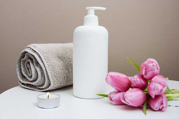 White pump bottle with a rolled towel, tealight candle, and pink tulips on a white surface, suggesting a spa or self-care setting
