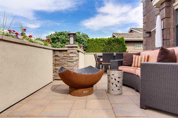 Outdoor patio with a rust-colored fire pit, wicker seating, and cushioned lounge chairs on tan tile, blue sky overhead.