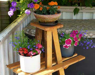 Wooden tiered plant stand on a porch with three pots of bright flowers: purple petunias, orange marigolds, and pink geraniums.