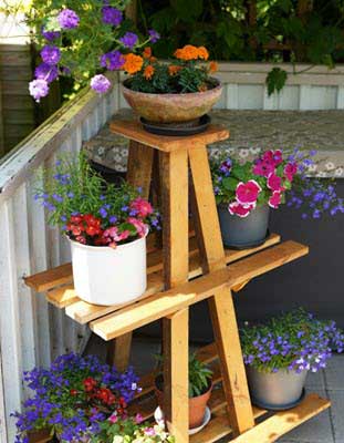 Wooden three-tier plant stand holding several colorful potted flowers on a balcony.