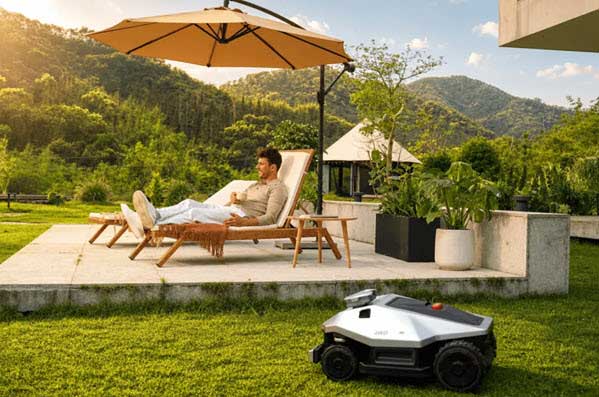robot lawmower small yards | Gardens with Wings Man relaxing on a lounge chair under a patio umbrella while a robotic lawn mower roams in the foreground on a sunny garden patio.