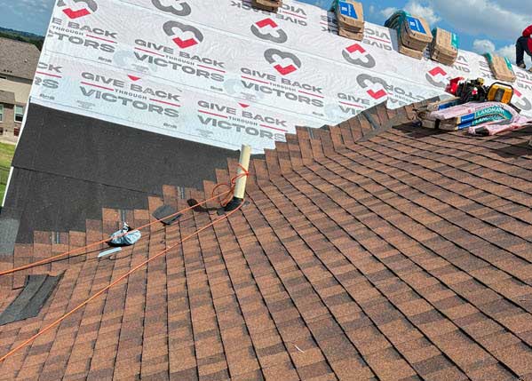 roof installation | Gardens with Wings Sloped roof under construction with brown shingles, a vent pipe protruding through, and construction material at the top edge against a white weather barrier with branding; an orange extension cord runs along the roof.
