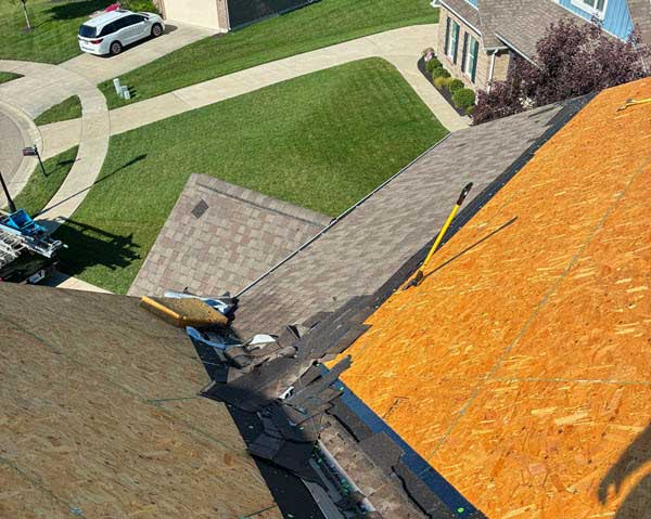 Rooftop under construction with orange plywood sheathing and dark underlayment, viewed from above; green lawn and houses visible in the background.