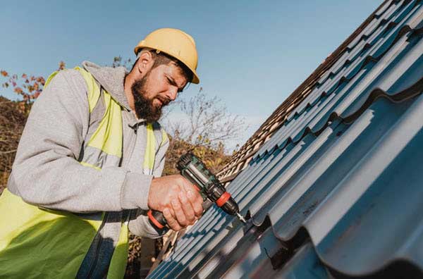 roofer | Gardens with Wings Construction worker in a yellow hard hat and high-visibility vest drilling into a metal roof panels at a sunny outdoor site.