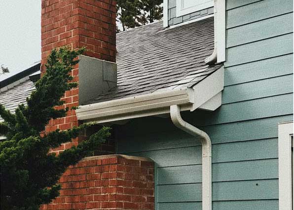 siding | Gardens with Wings Close-up of a house corner showing a brick chimney, blue siding, and white rain gutter with downspout along the eave.