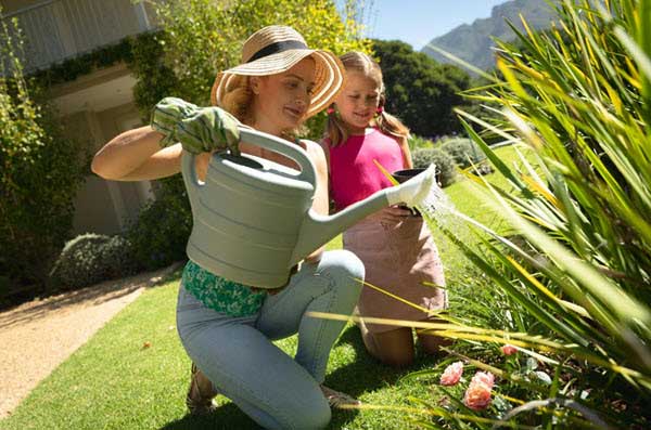 A women and child gardening in a sunny yard; one kneels, watering flowers with a large green watering can while the other watches.