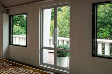 Interior of a room with a glass door and two side windows opening onto a balcony, flooding the space with natural light; pebbled floor along the wall.